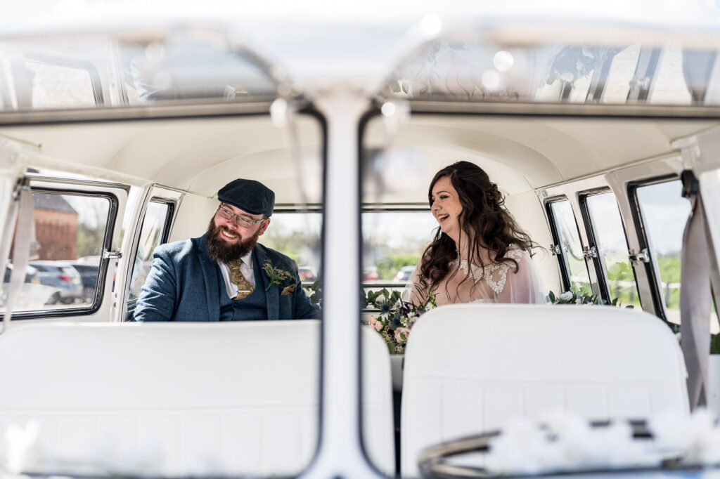 A bride and groom are joyfully interacting inside a vintage van, with the groom seated on the left wearing a blue suit and flat cap, and the bride on the right in a white dress with lace details. The van interior is white, and there's a view of the sunny outdoors through the windows.
